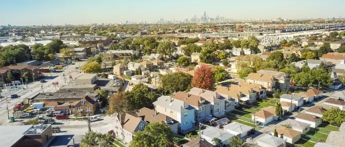 Aerial view of a Northeastern Illinois suburban neighborhood with single-family homes and tree-lined streets, with the Chicago skyline visible in the distant background.